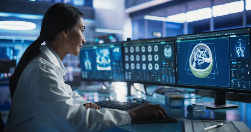 Female Asian Neurologist Using Desktop Computer With Human Brain MRI Scans In Modern Medical Research Center Laboratory.