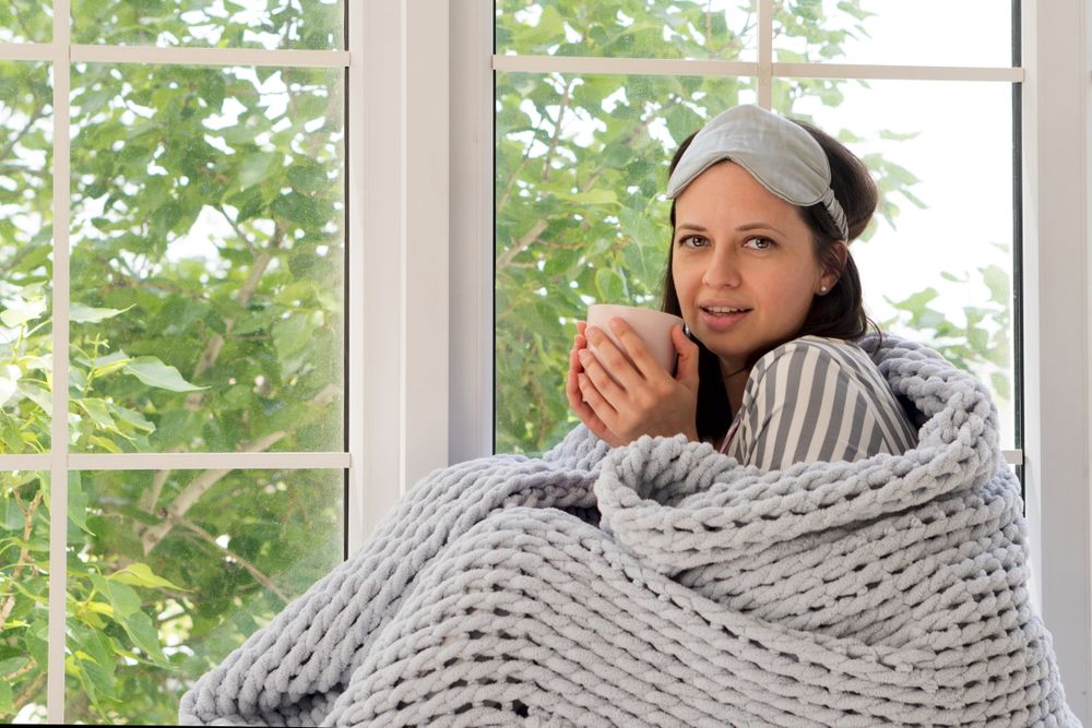Smiling woman wrapped in chunky blanket, holding mug near window.