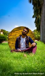 Man in traditional outfit sitting on green grass beside wooden spool bGBKY4