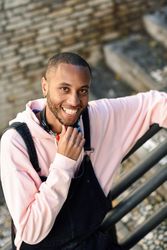 Happy male in overalls and hoodie leaning on railing of stairs outside, vertical