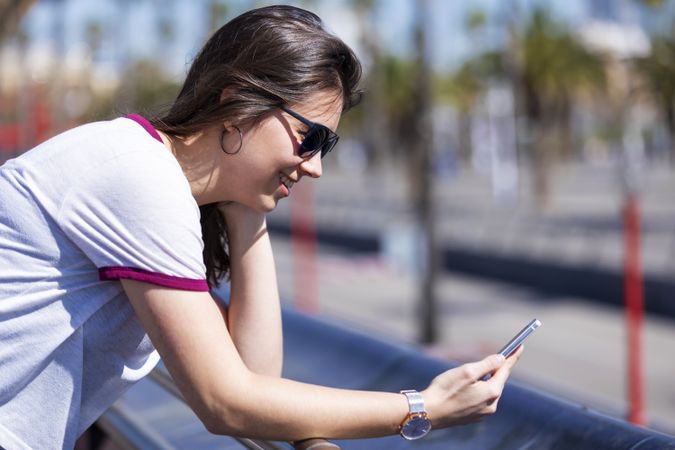 Side view of smiling woman wearing clothes standing outdoors in the street while checking phone on a sunny day