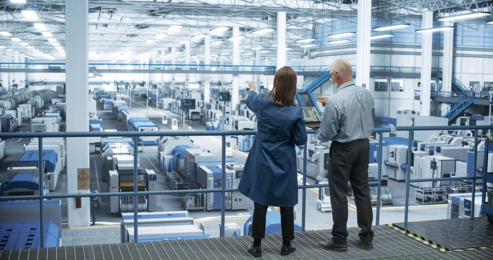 Male and Female Engineers Standing with Their Back to Camera, Using Laptop Computer and Talking in a Factory Facility with Equipment Producing Modern Electronic Components for Different Industries