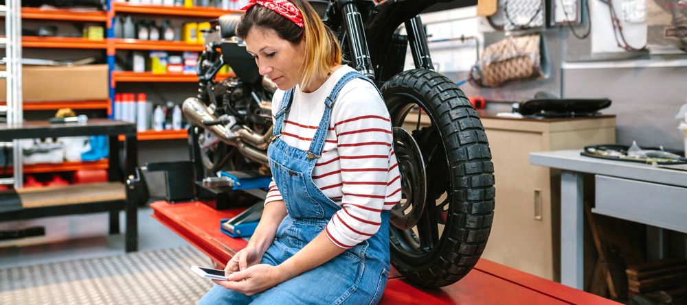 Mechanic woman looking phone sitting over platform with motorcycle on factory