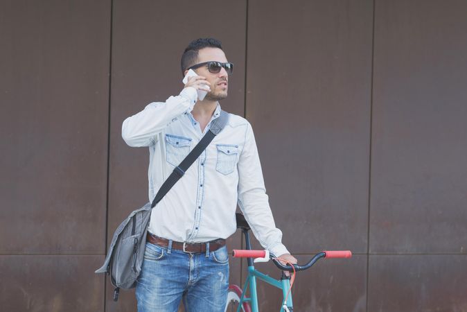 Male in sunglasses standing with red and green bicycle and looking around on phone
