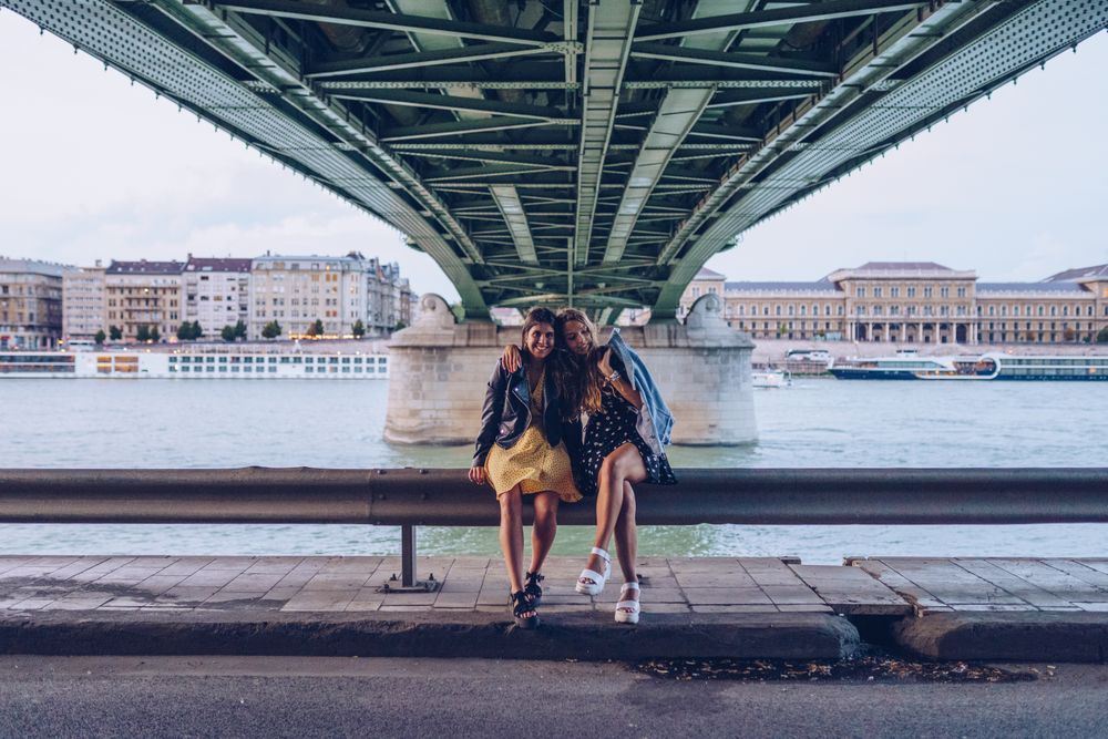 Smiling girlfriends hugging and sitting on fence along river under bridge