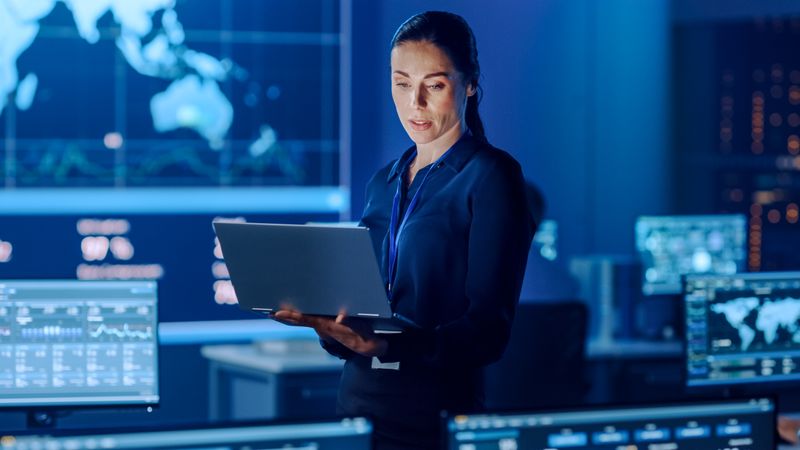 Focused Professional Woman Uses Laptop in Blue-lit Control Room. Surrounded by Data Screens and World Map, She Analyzes Information in High-tech Environment.