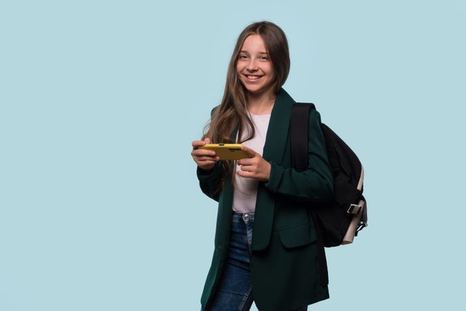 Teen girl smiling and holding phone, wearing backpack, casual look.