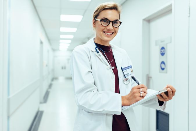 Portrait of smiling female doctor standing in hospital corridor with clipboard
