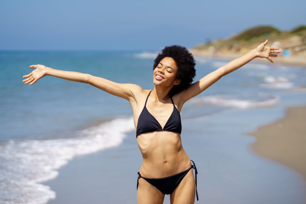 Happy woman in bikini standing on beach with outstretched arms and eyes closed