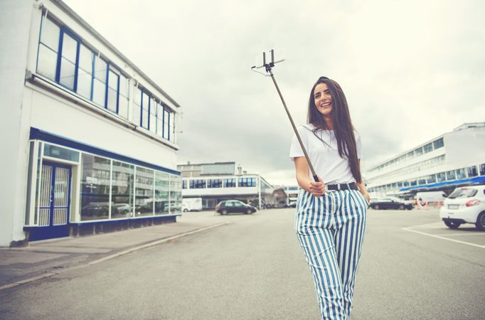 Woman with long brown hair posing in street with selfie stick for photo on overcast day