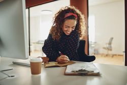 Woman with curly hair smiling while writing notes at her work computer