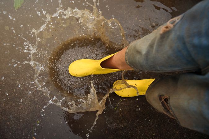 Crop person in rubber boots playing in puddle