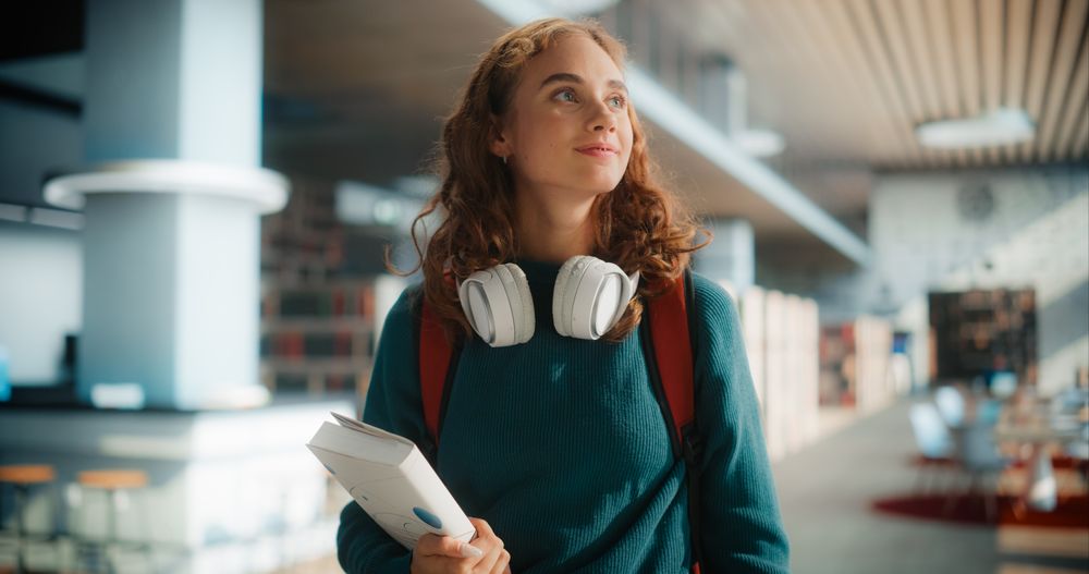 Dreamy Young Female Student Wandering Through Modern Library With Books and Headphones