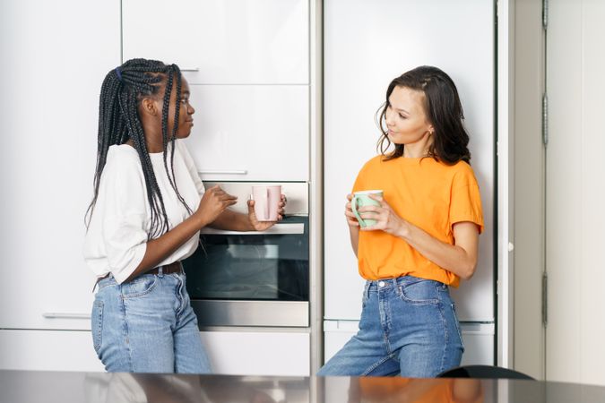 Two women talking while leaning on the fridge with coffee in hand
