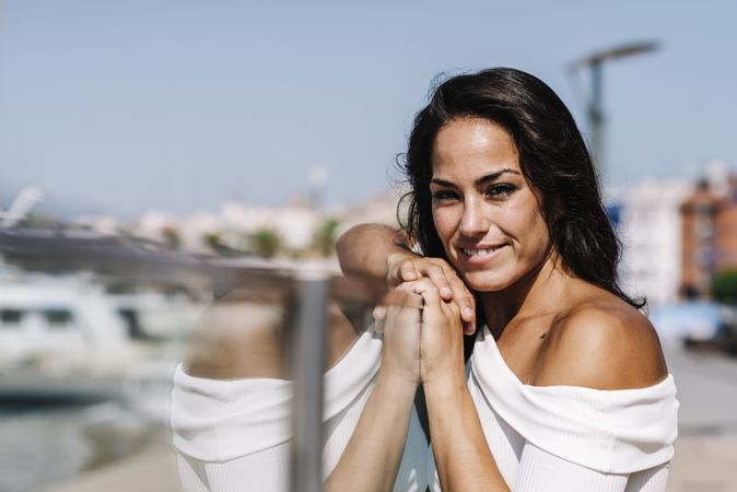 Front view of dreamy young woman leaning on fence while looking camera on harbor dock