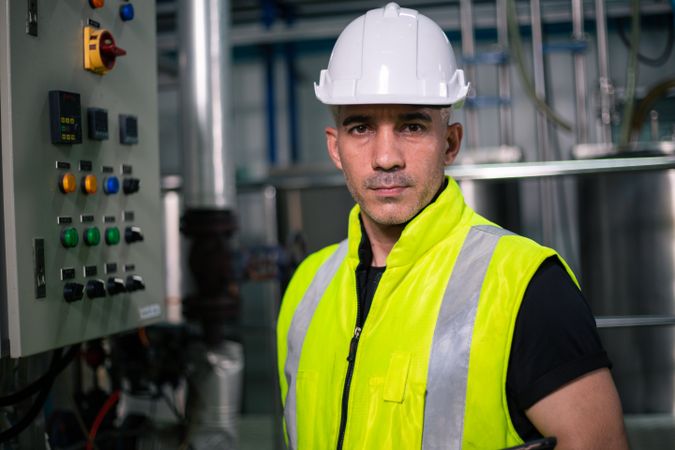 An industrial engineer wearing a safety helmet and reflective vest stands confidently in front of a machine in a factory. He is looking at the camera, representing professionalism and quality control