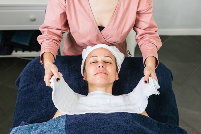 Crop cosmetologist steaming face of client with hot towel
