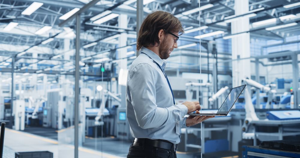 Experienced Male Engineer Standing in Office, Using Laptop Computer and Overlooking Production at Modern Automated Electronics Assembly Line with AI Support. Factory Executive at Work
