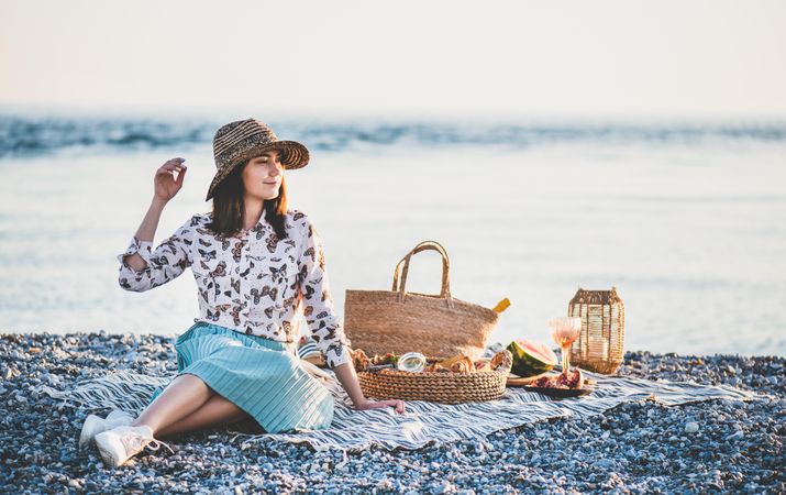 Young woman in hat sitting by the ocean with a picnic, watermelon, and wine