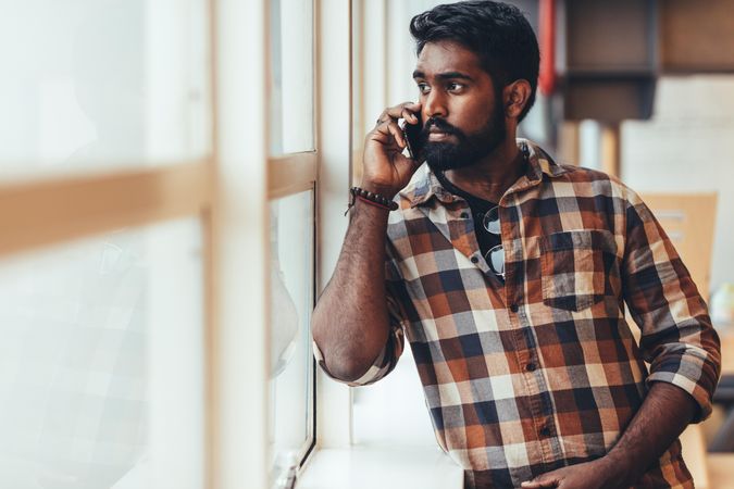 Indian male talking on smartphone standing beside a window