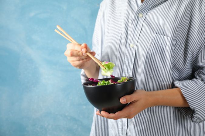 Young woman eat salad with chopsticks on blue background, space for text