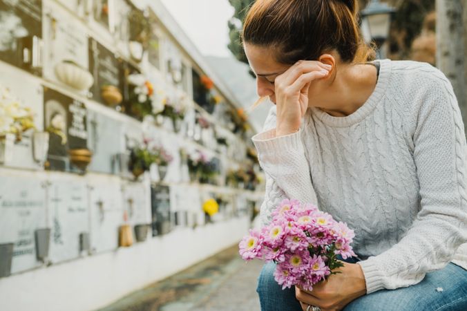 Woman with flowers crying near columbarium wall in cemetery