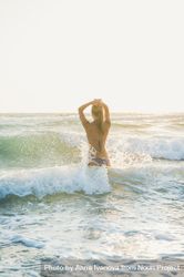 Rear shot of woman in bikini in waves at the beach with hands to her head 5lgn70