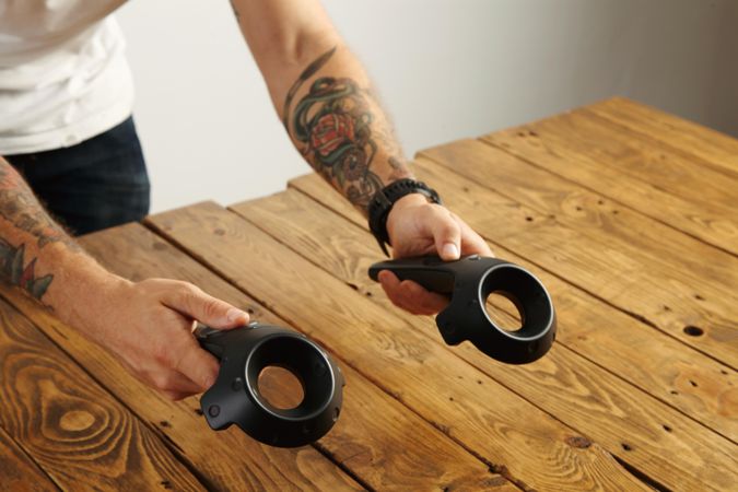 Tattooed hands hold two virtual reality remote controllers above wooden table