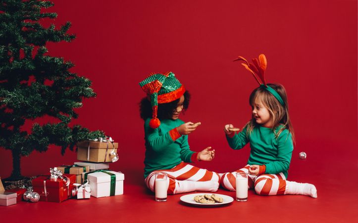 Little girls sitting beside a Christmas tree enjoying cookies and milk