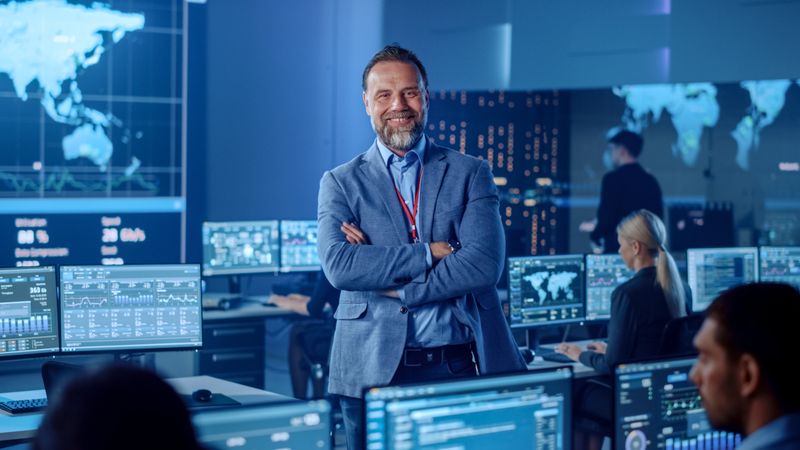 Confident Executive Stands in High-tech Control Room. Team Monitors Global Data on Screens. Blue-lit Environment With World Map Displays. Advanced Technology for Real-time Analysis.