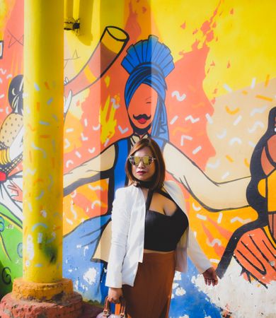 Woman in brown skirt and light blazer with sunglasses standing beside wall with graffiti