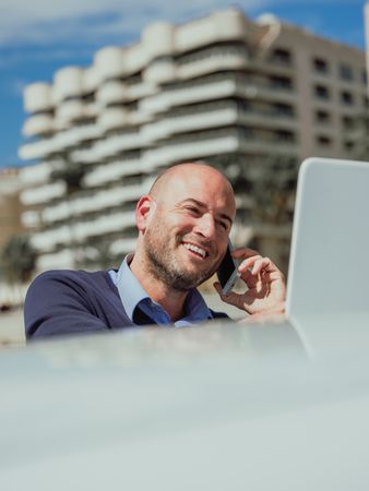 Businessman talking on the phone while using the laptop supported by a car with the buildings of the city behind