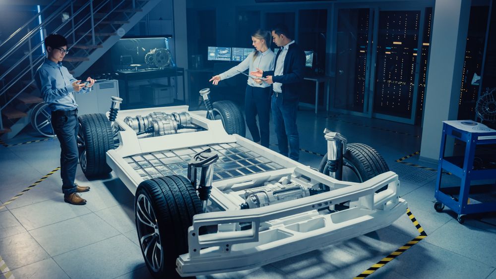 High Angle Shot of a Development Laboratory Room with Diverse Professional Team of Engineers and Scientists Working on a Electric Car Chassis with Wheels, Batteries, Engine and Suspension.