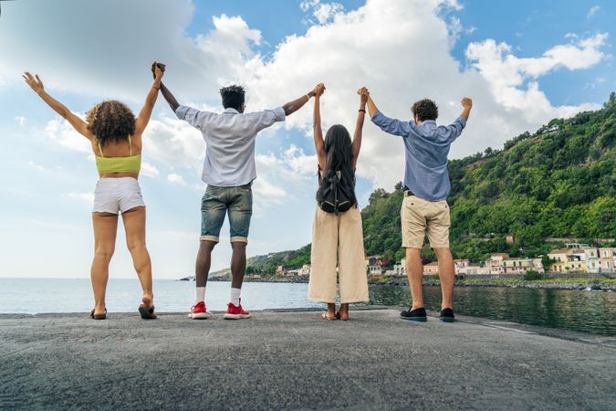 Back view of a group of diverse friends holding hands with arms raised, celebrating on a pier by the sea.