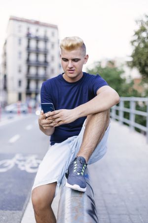 Man with blonde hair sitting on the outdoor railings and using a mobile phone