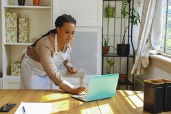 Female entrepreneur in work apron using laptop in bright office