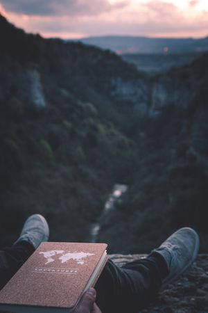 View to small river in canyon and legs of person with book sitting on the edge