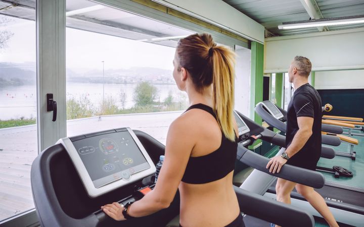 Woman and man on treadmills in gym overlooking lake view from window