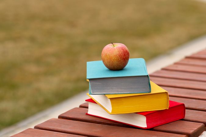 Single apple on stack of books placed outdoors bench.