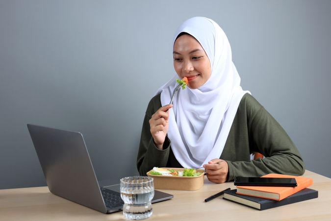 Smiling Woman Wearing Hijab Eating Healthy Salad While Working on Laptop.