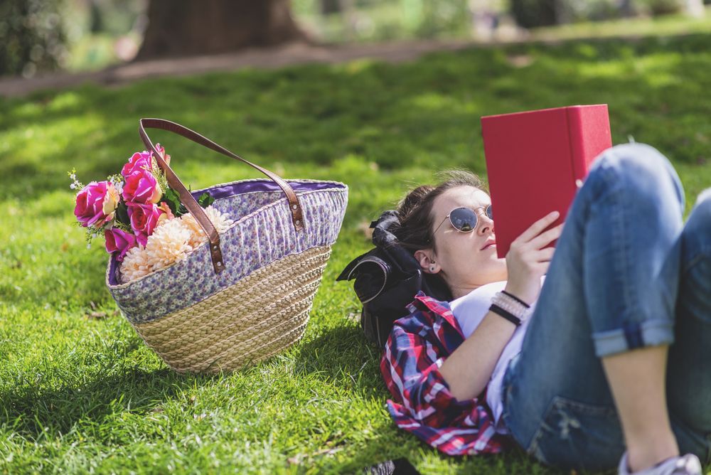 Side view of a hipster happy woman lying on grass in sunny day at park while reading a red book