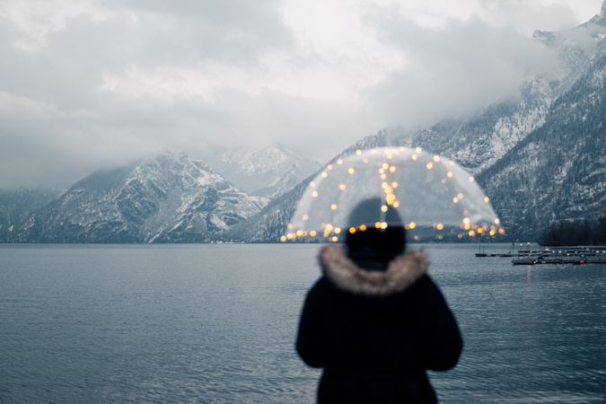 Back view of person holding an umbrella standing against body of water and snow covered mountainous landform in Slovenia