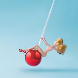 Barbie like doll swinging on red festive bauble on blue background