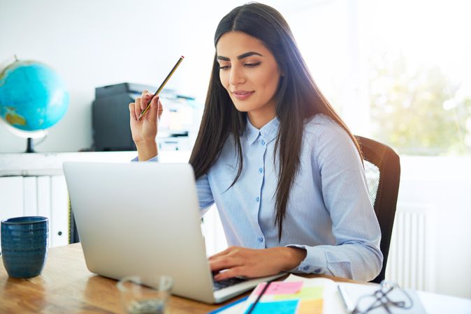 Woman working on laptop at hope in bright office while holding pencil