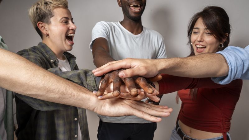 Team of diverse young adults joining hands in teamwork gesture, laughing and smiling together