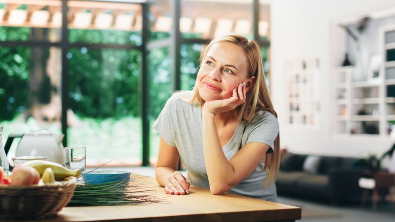 Portrait of Young Adult Woman with Blond Hair Wearing Gray V-Neck T-Shirt, Leaning on a Table and Smiling