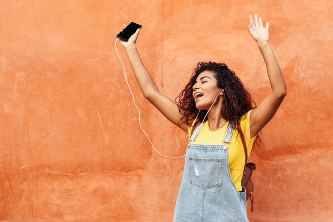 Dancing Arab woman with curly hairstyle using phone and headphones with arms in the air