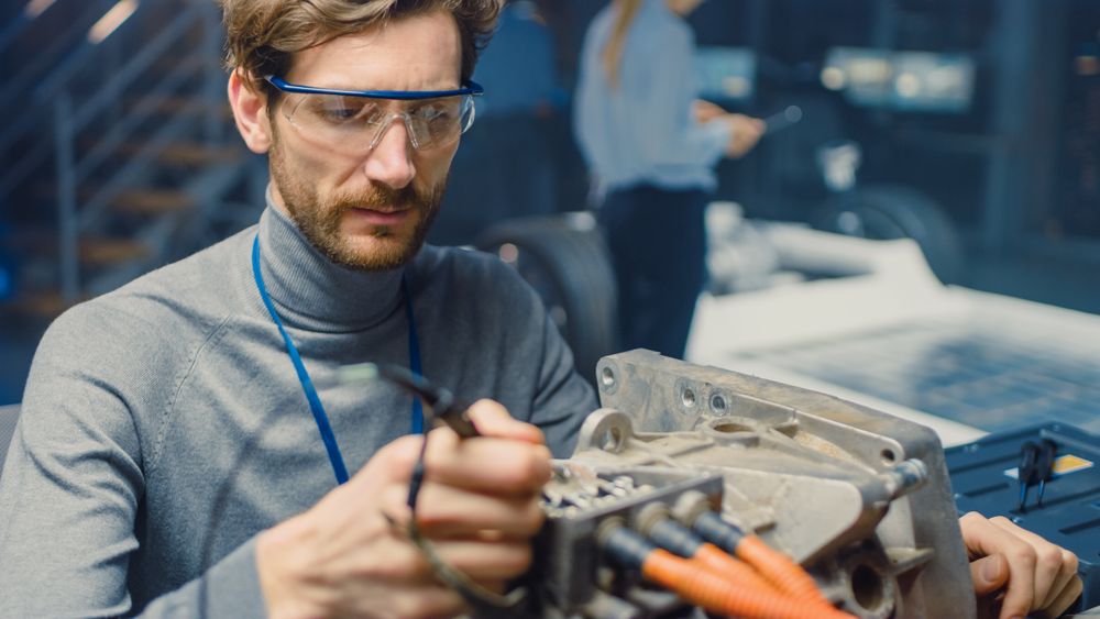 Professional Automotive Engineer in Glasses with a Computer and Inspection Tools is Testing an Used Electric Engine in a High Tech Laboratory with a Concept Car Chassis.