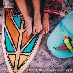 Women’s feet resting atop a patterned surf board in the sand B5QBX4