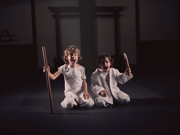 Funny kids in judogi sitting on mat with wooden sticks and shouting